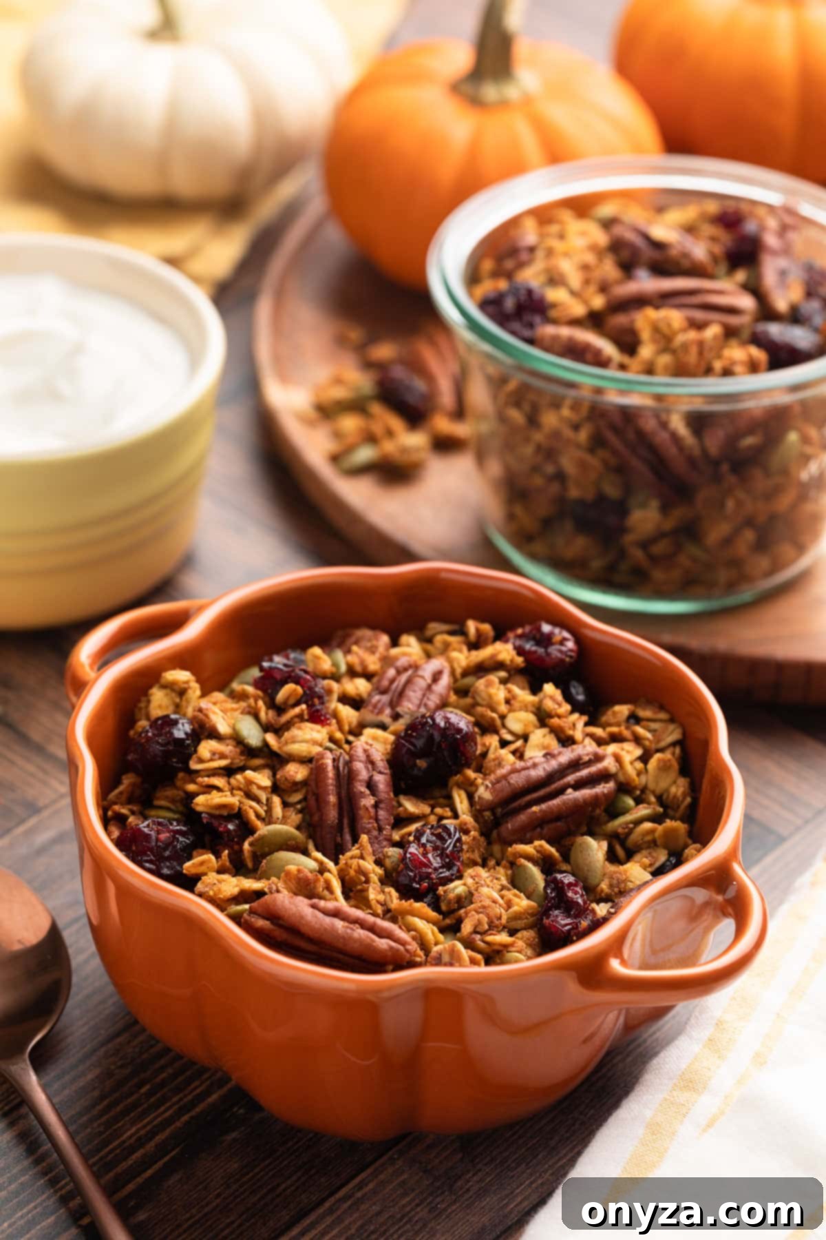 Pumpkin granola in a pumpkin cocotte. In the background is a glass jar of granola, a bowl of yogurt, and miniature pumpkins, evoking a cozy fall atmosphere.