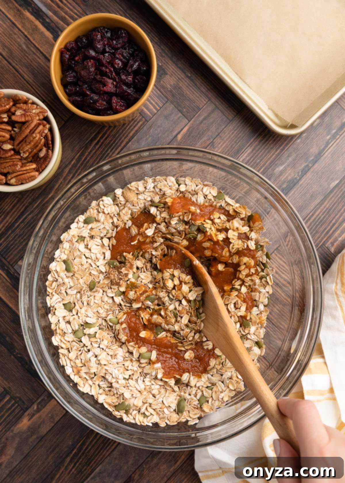 Overhead photo of the wet pumpkin granola ingredients being gently stirred into the dry ingredients in a clear glass bowl with a wooden spoon, ensuring an even coating.