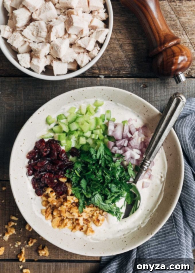 A bowl of chicken salad dressing with dried cranberries, diced celery, chopped shallots, fresh parsley, and toasted walnuts, ready to be mixed.