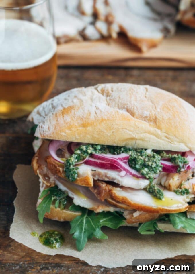 A delectable Porchetta Sandwich featuring thinly sliced porchetta, vibrant Italian salsa verde, and crisp pickled onions, all nestled within a toasted ciabatta roll, presented on a rustic wooden board.