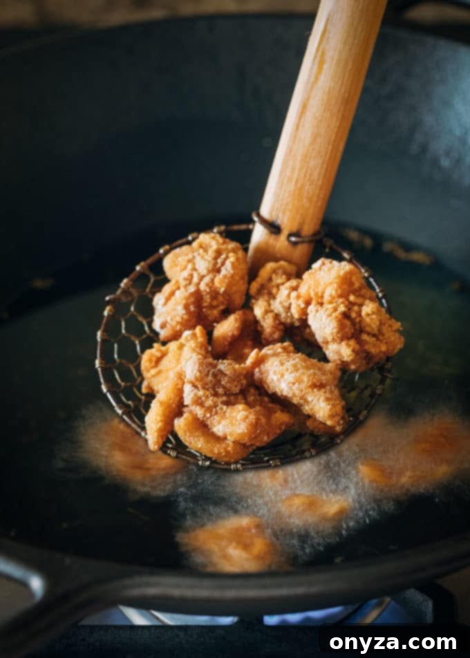Golden-brown, crispy fried chicken thigh pieces being lifted from hot oil with a spider strainer, ready to be drained before saucing