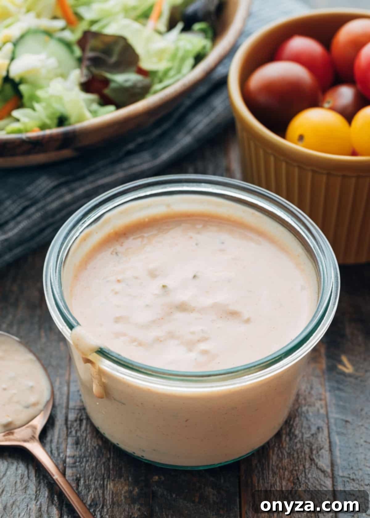 Homemade Russian dressing in a glass jar next to a bowl of cherry tomatoes and a vibrant green salad