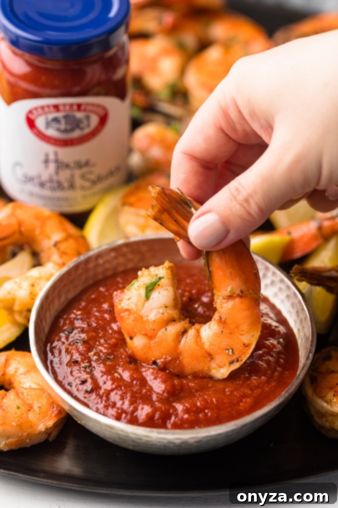 Signature Roasted Shrimp with Duo Dips 5 A perfectly roasted colossal shrimp being dipped into a rich, zesty bowl of Bloody Mary cocktail sauce, highlighting its vibrant color and succulent texture.