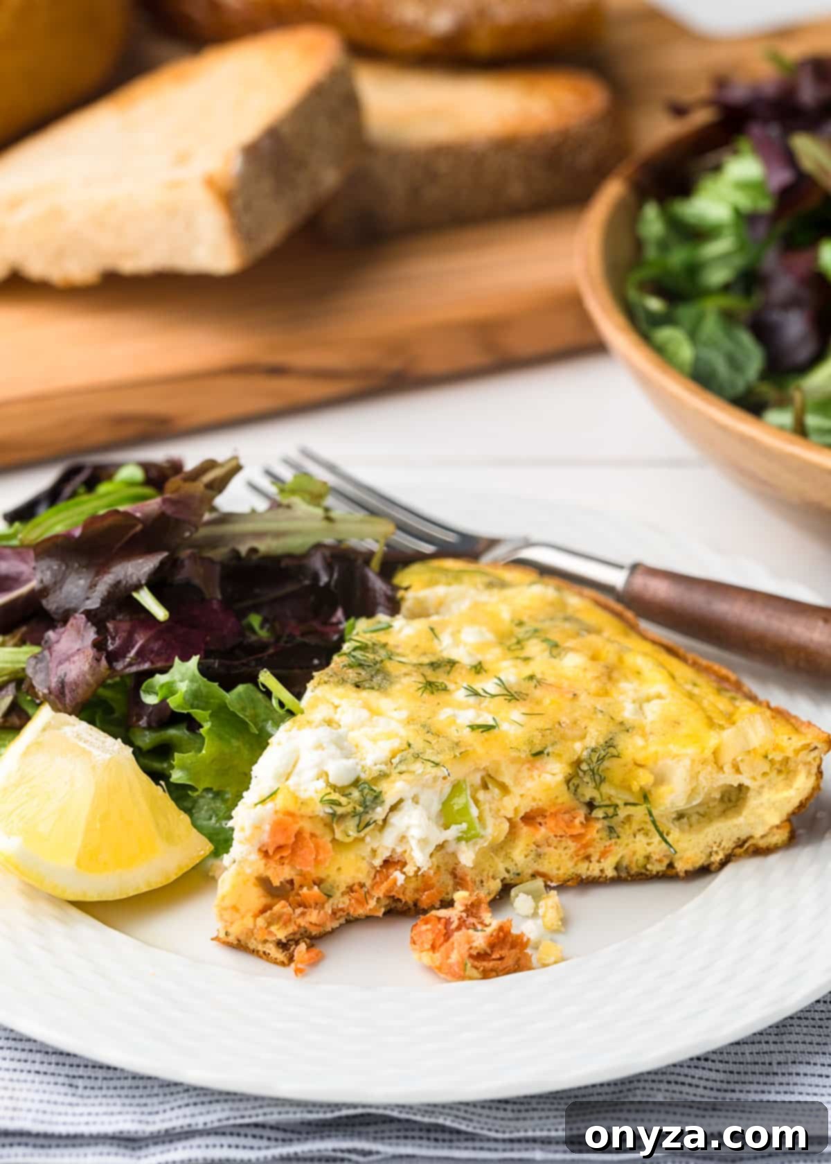 slice of smoked salmon frittata on a white plate with spring greens and a lemon wedge
