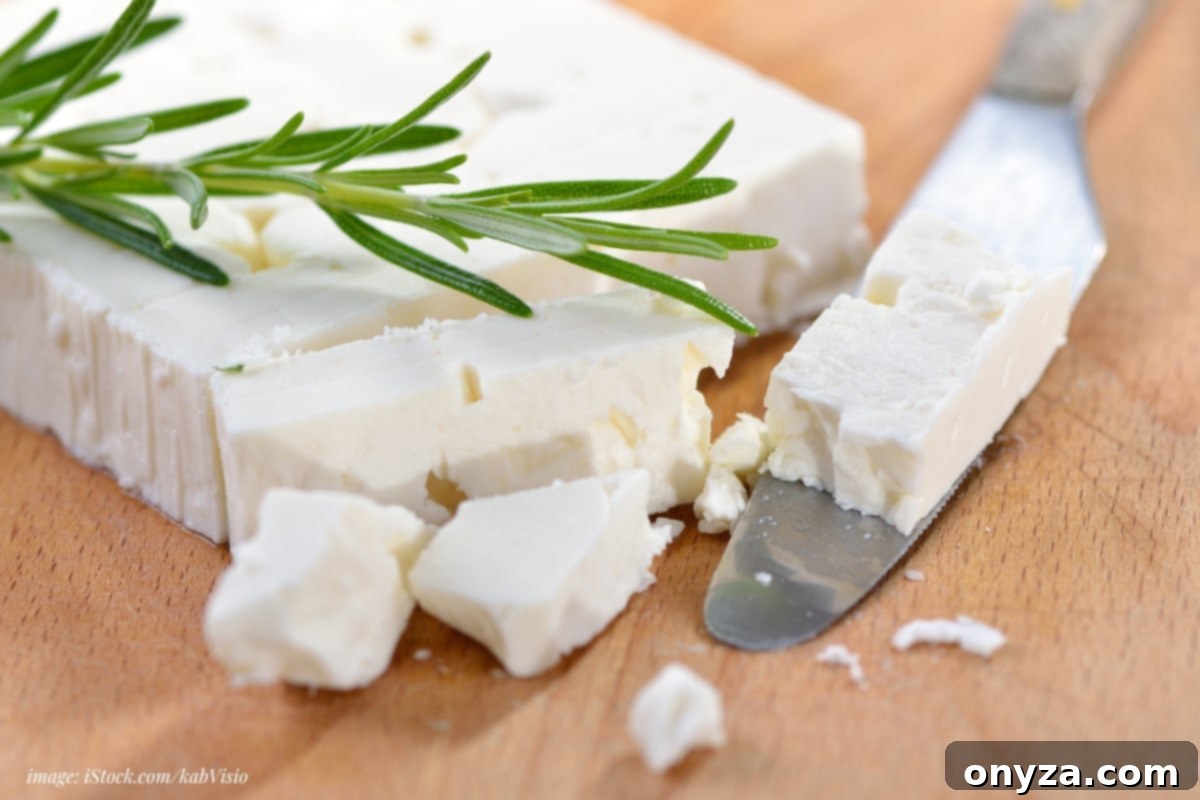 feta block on a wood board, partially crumbled with a cheese knife and sprig of rosemary
