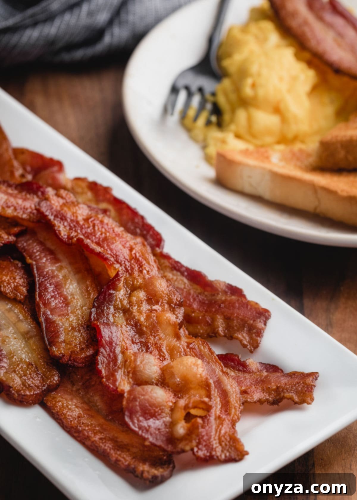 Perfectly baked bacon strips served on a white platter alongside scrambled eggs and toast, ready for breakfast.
