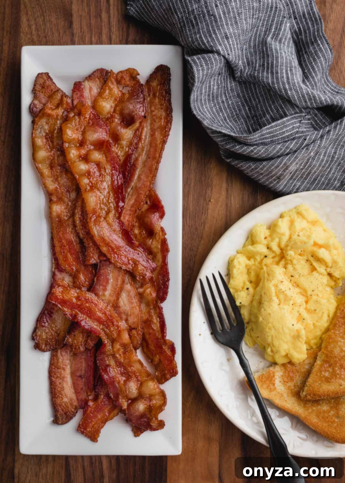 Overhead view of crispy baked bacon arranged artfully on a white rectangular platter, with a breakfast plate of eggs nestled beside it.