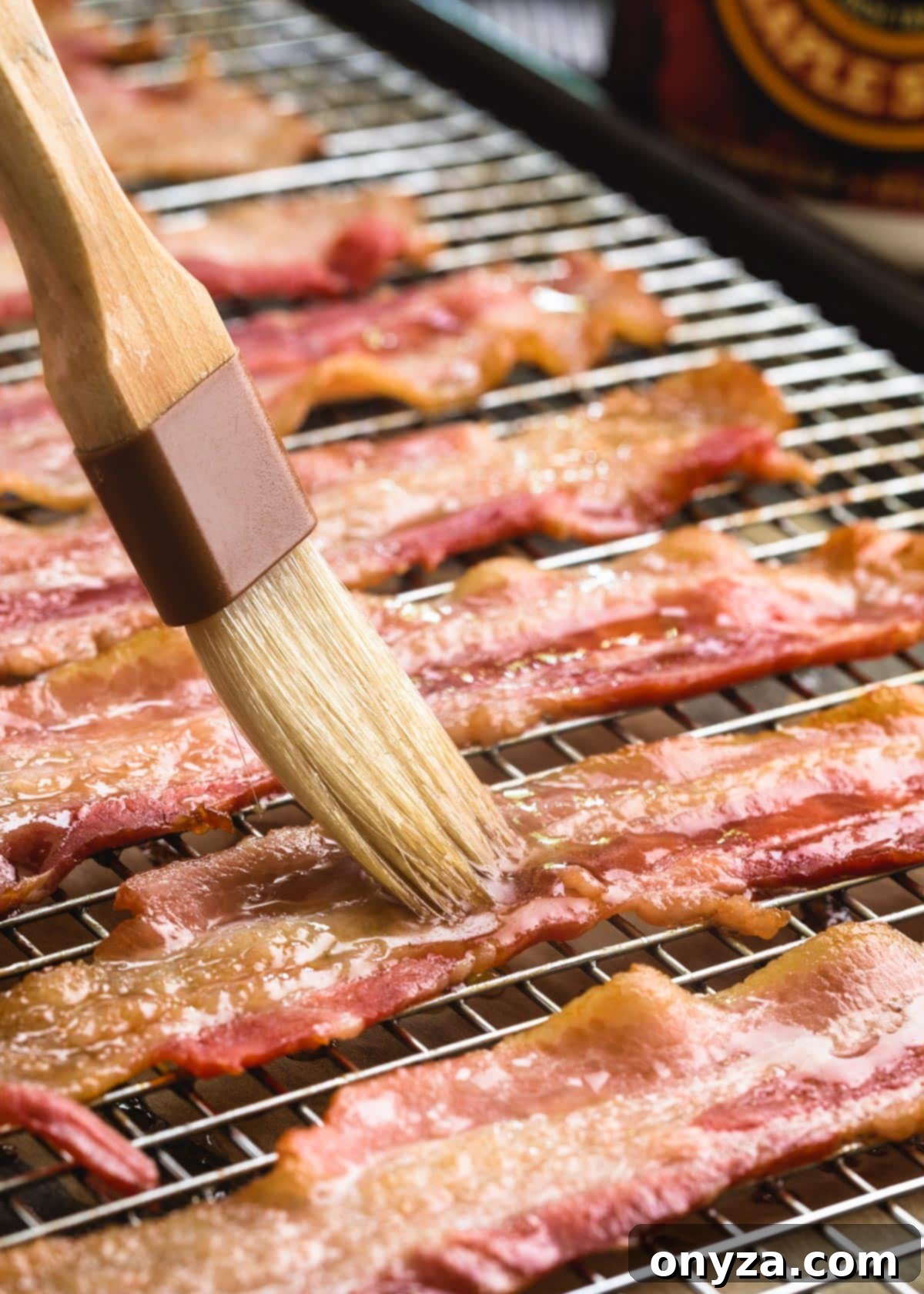 Maple syrup being delicately brushed onto partially-baked bacon strips arranged on a wire rack, adding a sweet glaze.