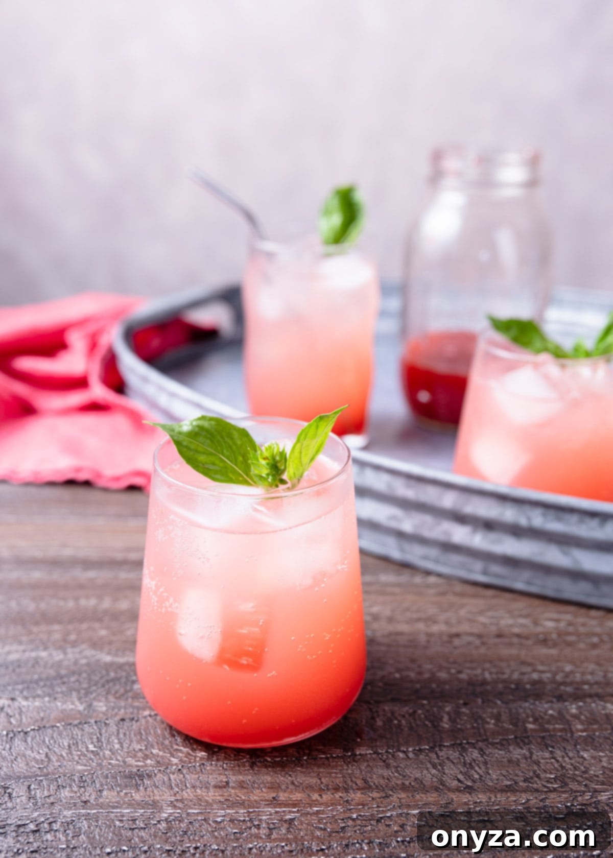 Several glasses of Strawberry Basil Soda, garnished with fresh basil and strawberry slices, are artfully arranged on a wooden board and a sleek metal serving tray.