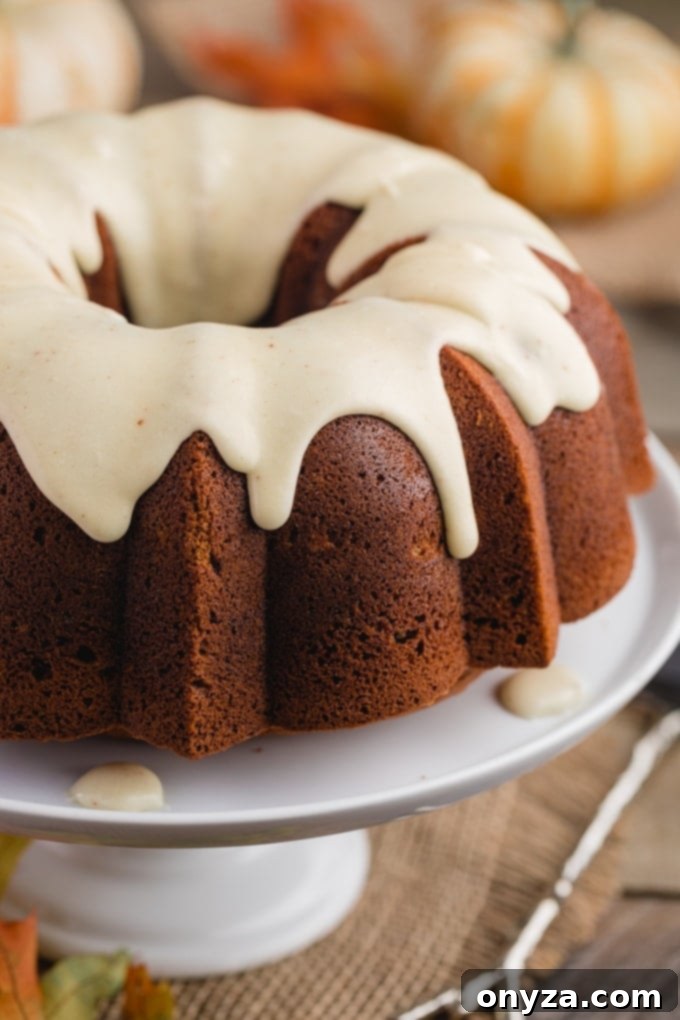 A beautifully glazed pumpkin bundt cake, presented on a white cake pedestal, surrounded by charming autumn decorations like mini pumpkins and colorful leaves, creating a festive display.