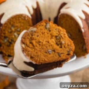 A slice of moist Pumpkin Bundt Cake on a cake server, with the full cake visible on a white pedestal in the background.