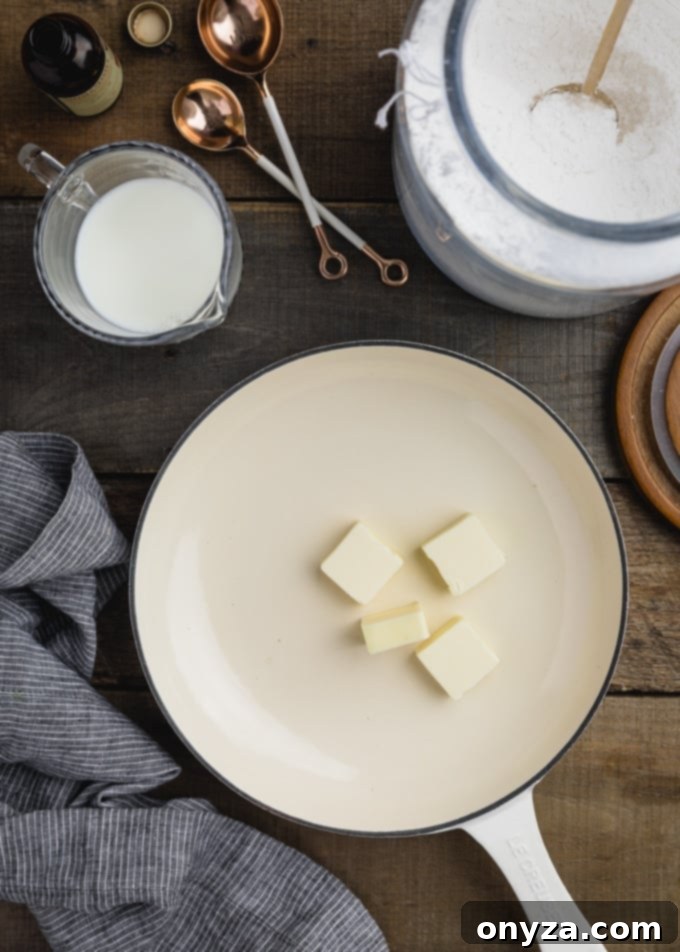 pieces of butter in a white skillet next to a pitcher of milk and canister of powdered sugar