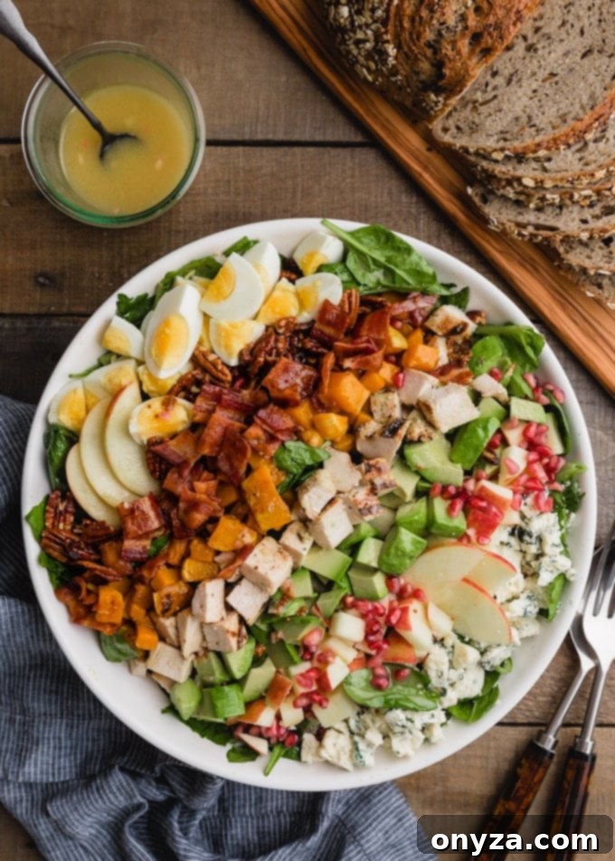 Overhead view of a beautifully arranged Harvest Cobb Salad in a white bowl, accompanied by a jar of maple-Dijon vinaigrette and slices of whole-grain bread.