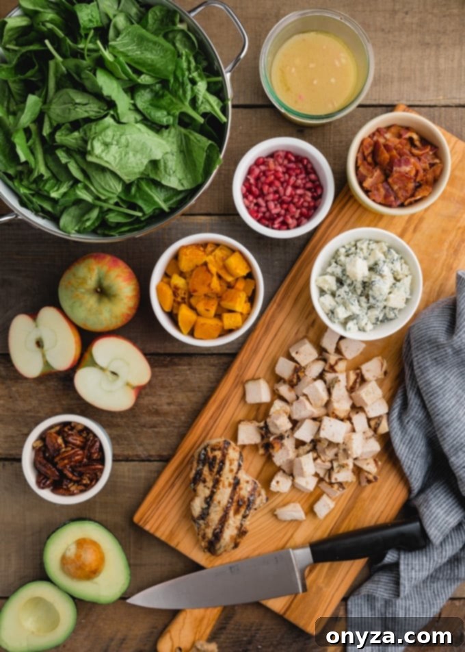 Ingredients for Harvest Cobb Salad laid out on a wooden board, including diced apples, cooked chicken, roasted butternut squash, mixed greens, and blue cheese crumbles.