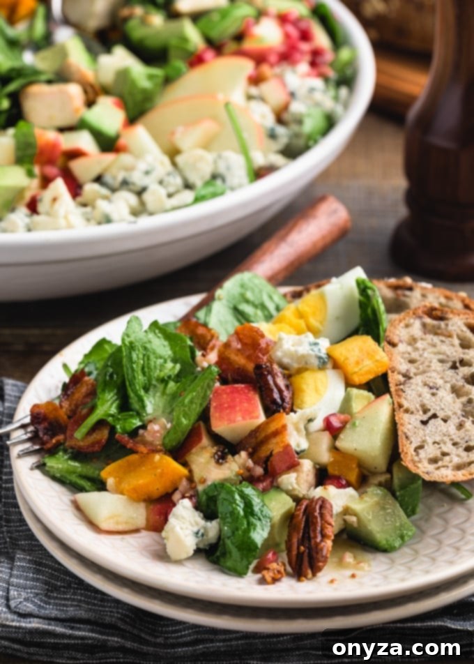 Side view of a vibrant Harvest Cobb Salad served on an ivory plate, featuring distinct rows of apples, pecans, butternut squash, and other ingredients, alongside a slice of whole grain bread.