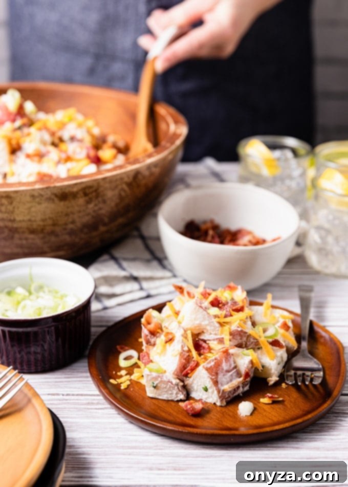 Delicious loaded potato salad with bacon and cheddar on a rustic wooden plate, surrounded by bowls of fresh scallions, crumbled bacon, and a large serving bowl, ready for a summer gathering.