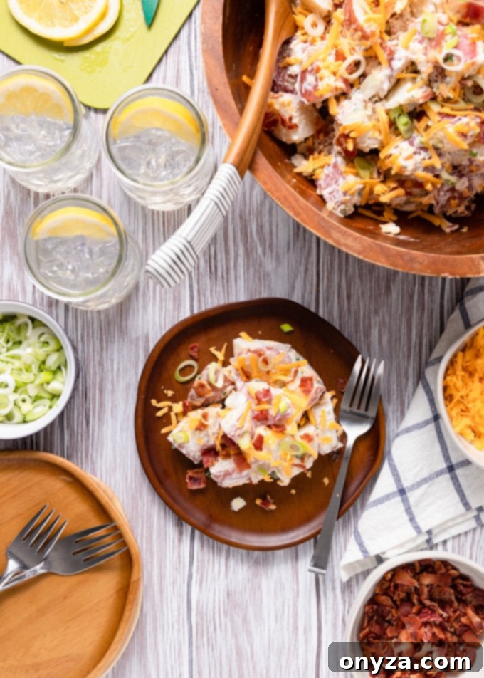 An inviting overhead view of a large wooden serving bowl filled with creamy bacon cheddar potato salad, with a smaller plate offering a portion and glasses of refreshing summer drinks in the background.