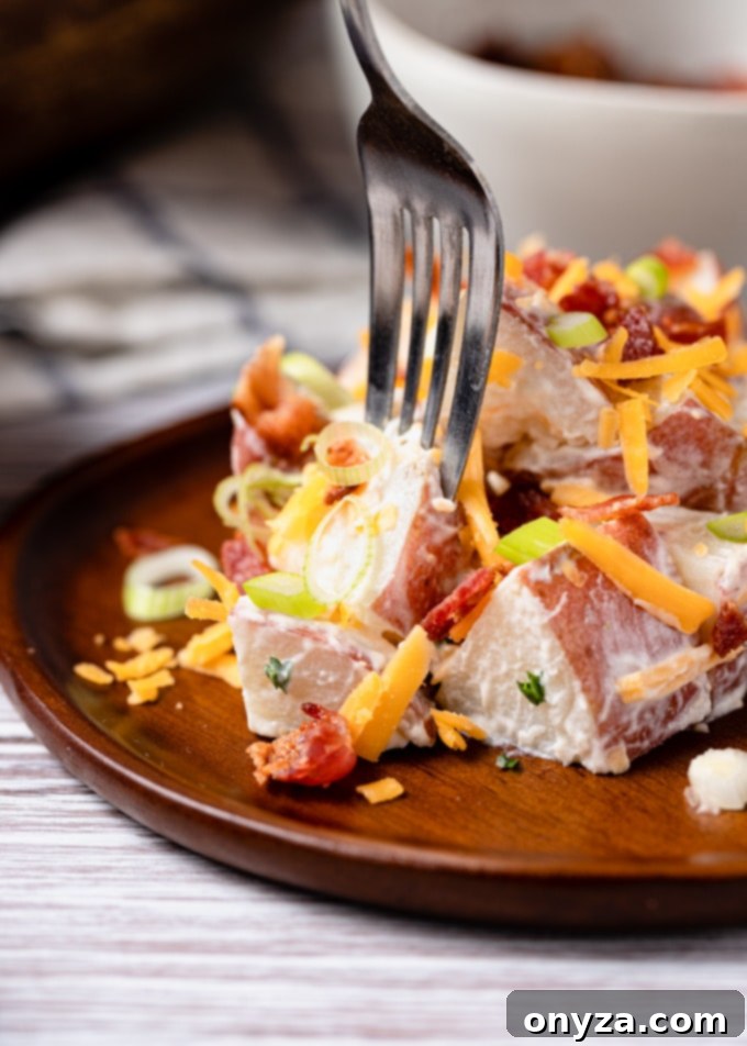 A close-up side view of a fork lifting a perfect, creamy bite of loaded potato salad from a wooden plate, showcasing its texture and flavorful ingredients.