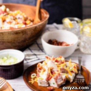 A beautifully presented serving of loaded potato salad on a wooden plate, with a large wooden serving bowl full of the salad in the background.