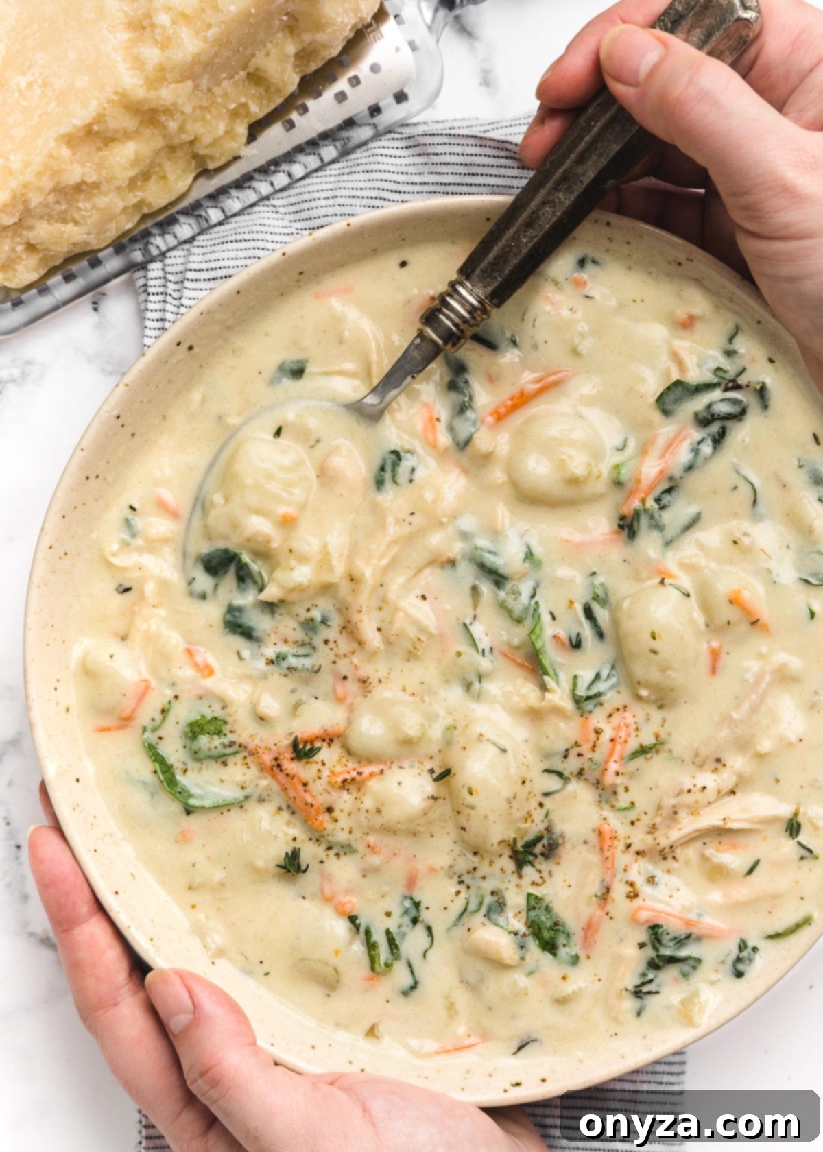 Hearty Chicken Gnocchi Comfort 13 overhead photo of two hands holding a bowl of creamy chicken gnocchi soup. One hand holds a silver spoon.