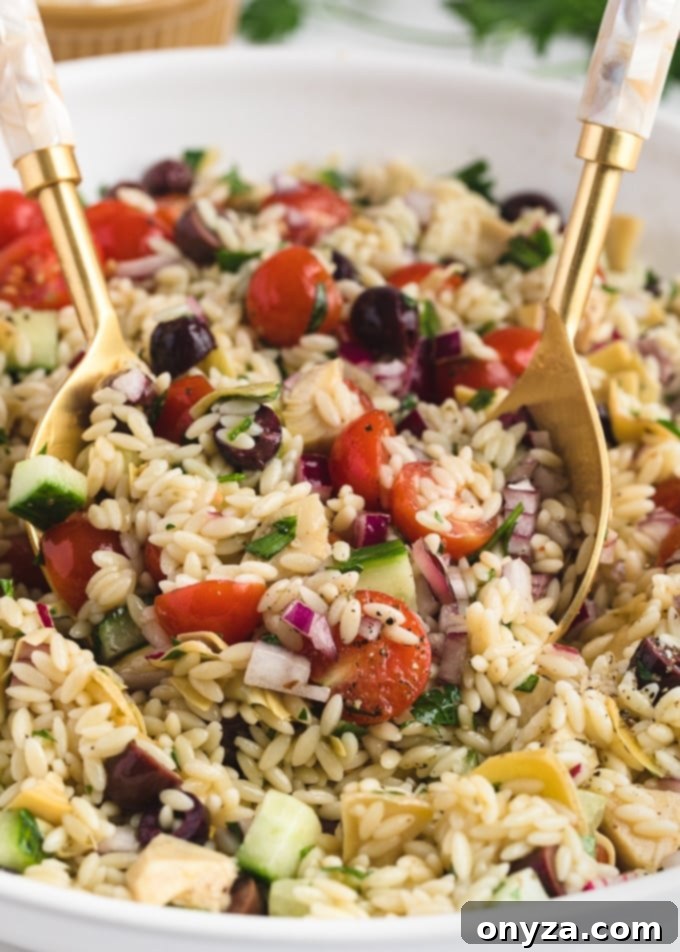 Close-up of Orzo Salad being tossed with a stylish gold salad spoon and fork set in a large white bowl, highlighting the mixed ingredients and vibrant colors.