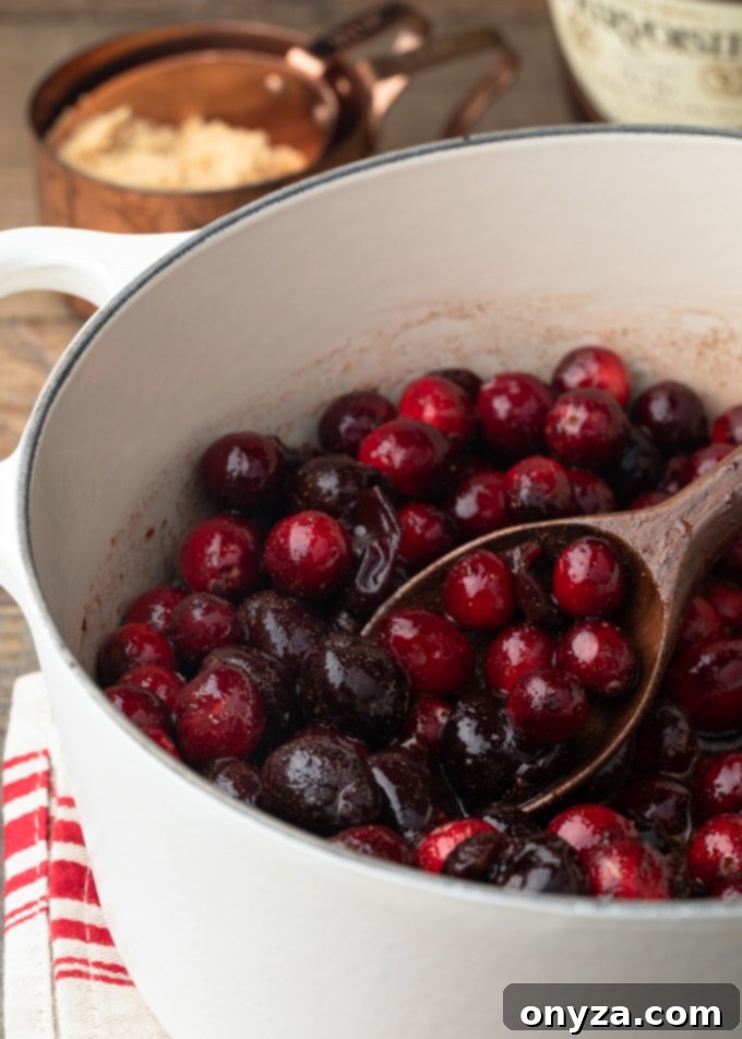 cranberries and frozen dark cherries starting to cook down in a white pot with a wooden spoon