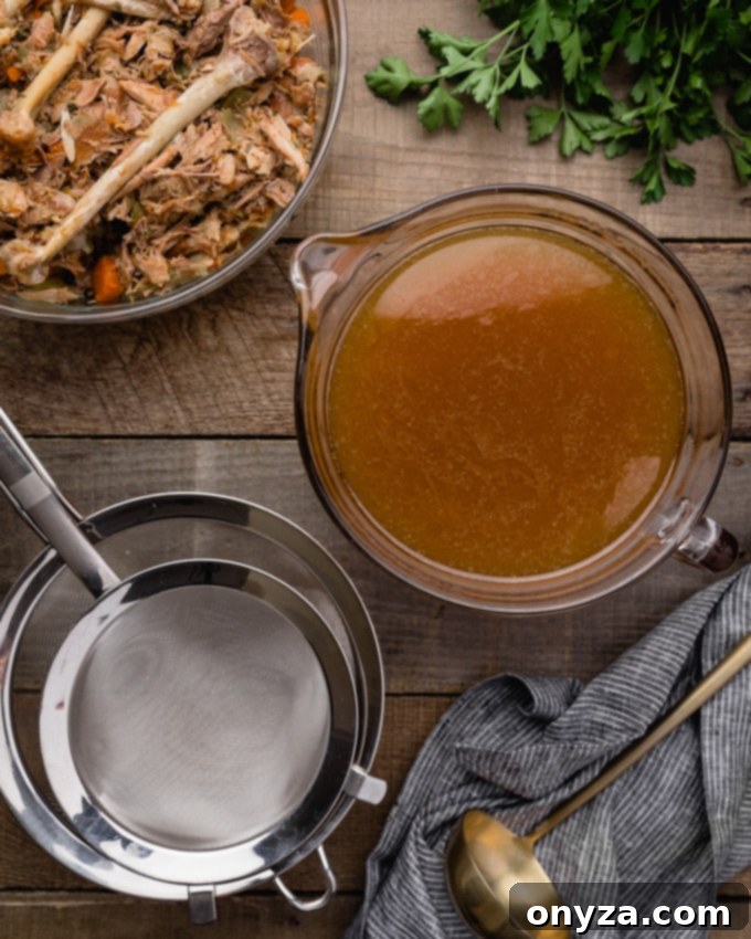 A bowl of strained, golden turkey stock next to discarded turkey meat, bones, and vegetables, with mesh strainers nearby.