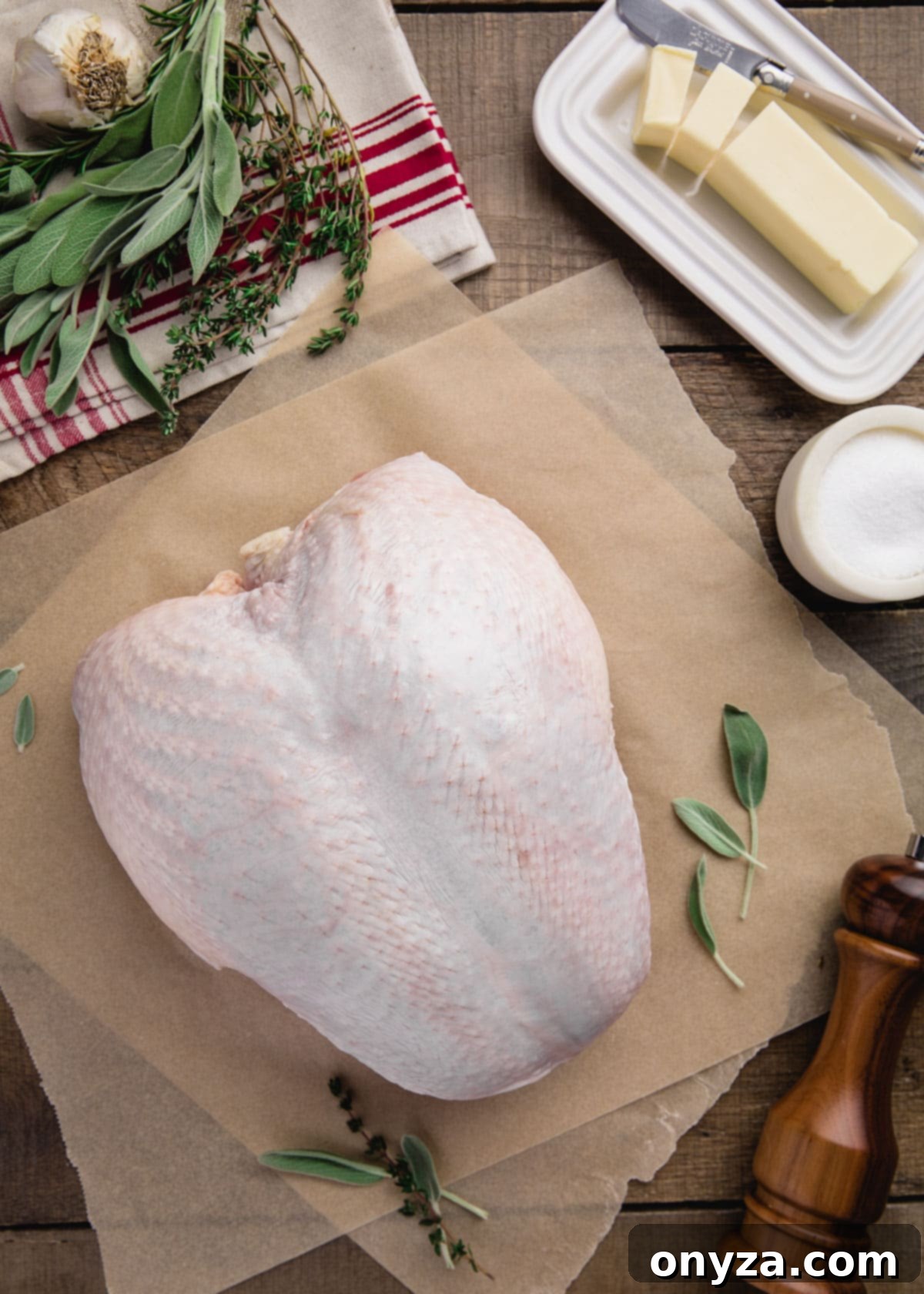 Raw whole turkey breast on parchment paper, surrounded by softened butter, fresh herbs, and various seasonings