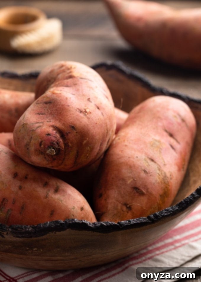 Velvety Brown Butter Sweet Potatoes 3 Raw, unpeeled sweet potatoes neatly arranged in a large wooden bowl, resting on a beige and red plaid napkin.