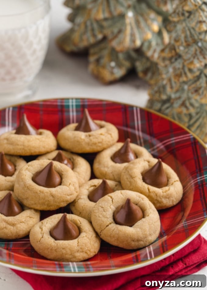 Peanut butter blossoms on a red plaid plate with a glass of milk and pine tree-shaped candles in the background, creating a cozy holiday atmosphere.