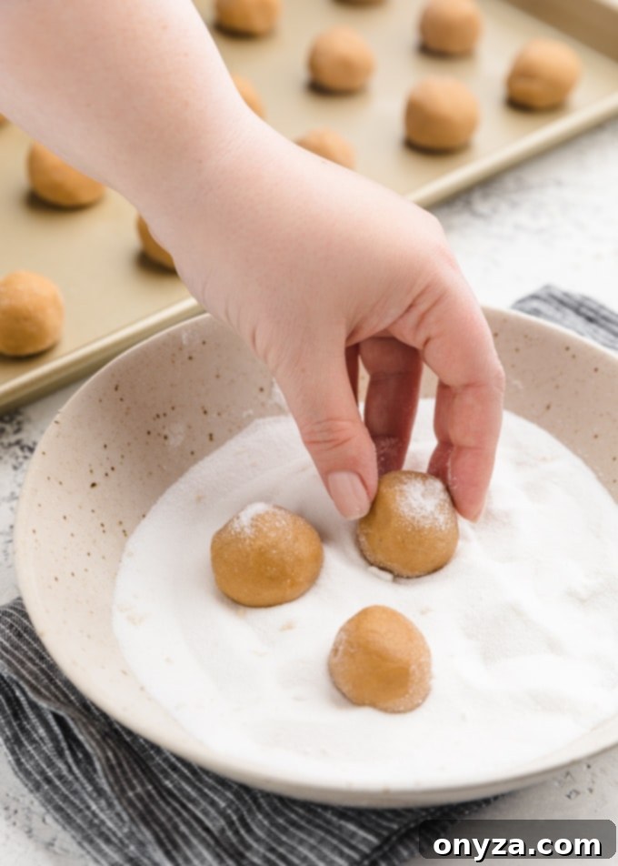 Rolling balls of peanut butter cookie dough in granulated sugar in a ceramic bowl, preparing them for baking.