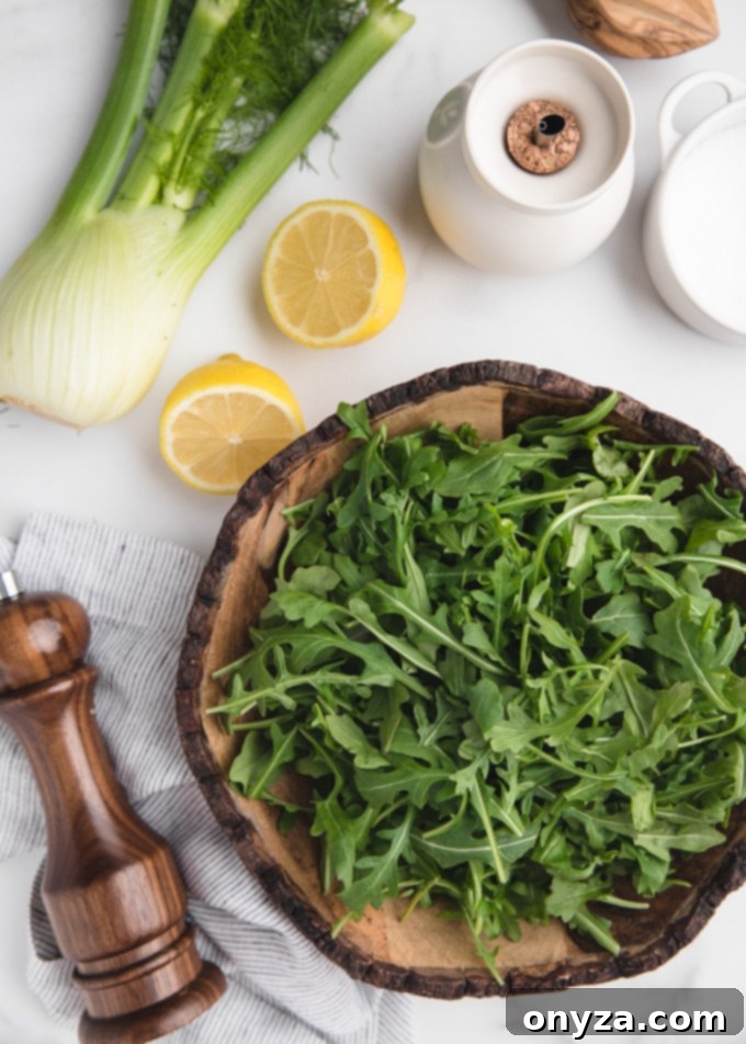 Overhead shot showcasing the fresh ingredients for an arugula salad: a whole bulb of fennel, bright lemon halves, a bowl brimming with baby arugula, a bottle of olive oil, and small dishes of salt and pepper, all arranged on a pristine white marble surface.