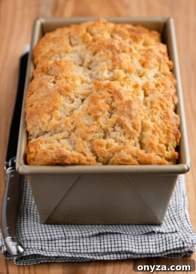 A baked loaf of beer bread in a gold loaf pan on a wooden board with a white and blue checked napkin