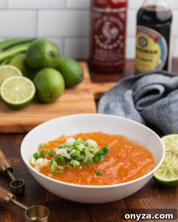 A bowl of bright orange marmalade is garnished with finely sliced scallions, surrounded by fresh whole and halved limes, along with bottles of soy sauce and Sriracha, highlighting the key ingredients for the dipping sauce.