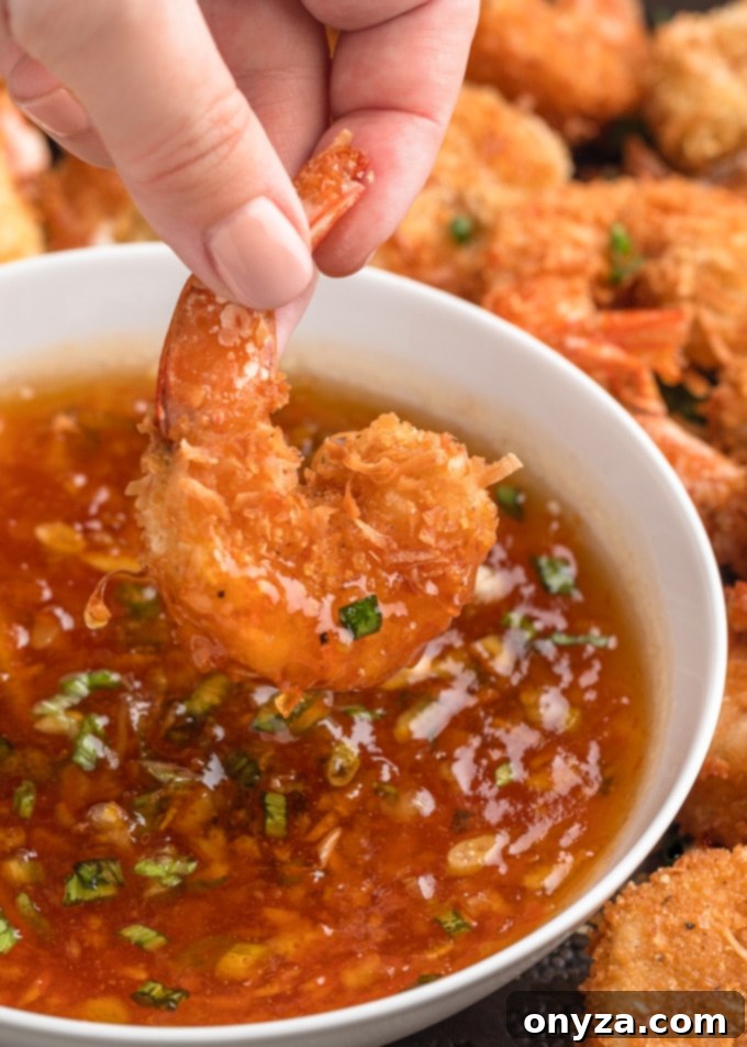A piece of golden-brown coconut shrimp is delicately dipped into a small white bowl of vibrant spicy orange dipping sauce, with more shrimp visible in the background.
