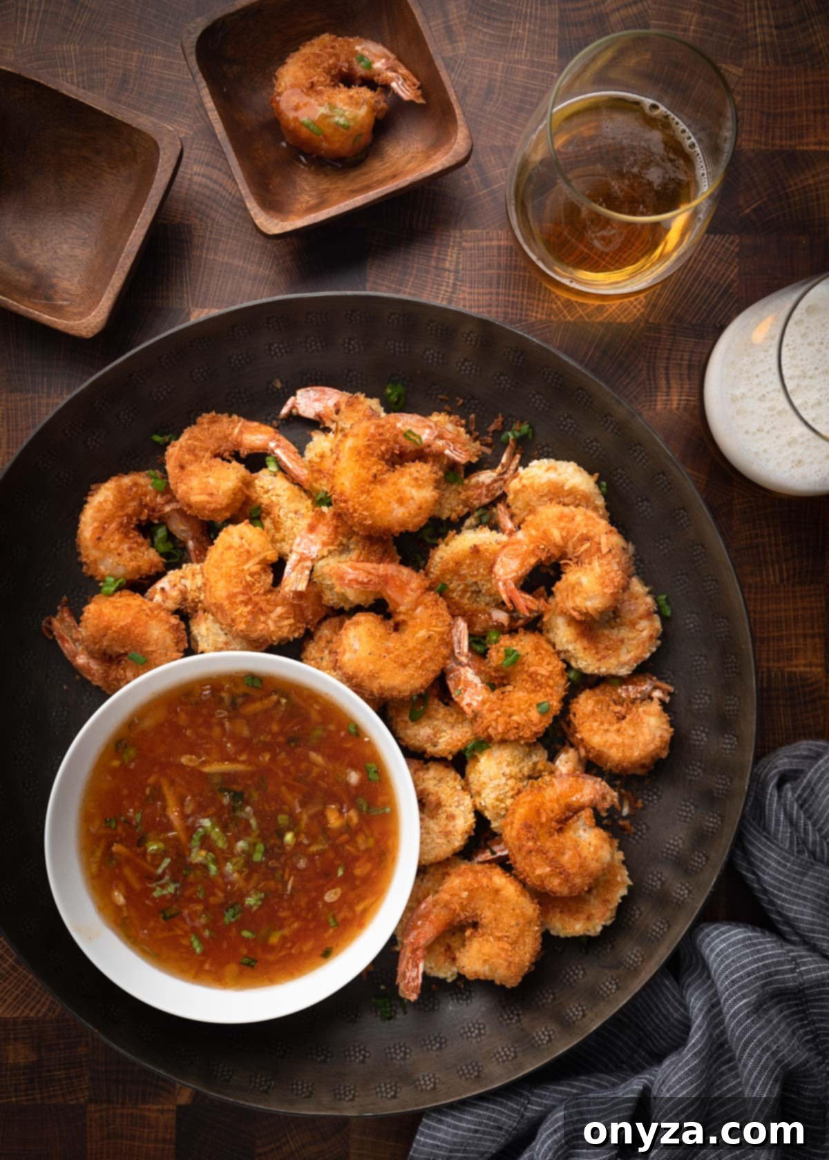 Tropical Crispy Prawns 2 Golden, crispy coconut shrimp arranged on a bronze serving platter, accompanied by a vibrant orange chili dipping sauce and two glasses of beer, creating an inviting appetizer display.