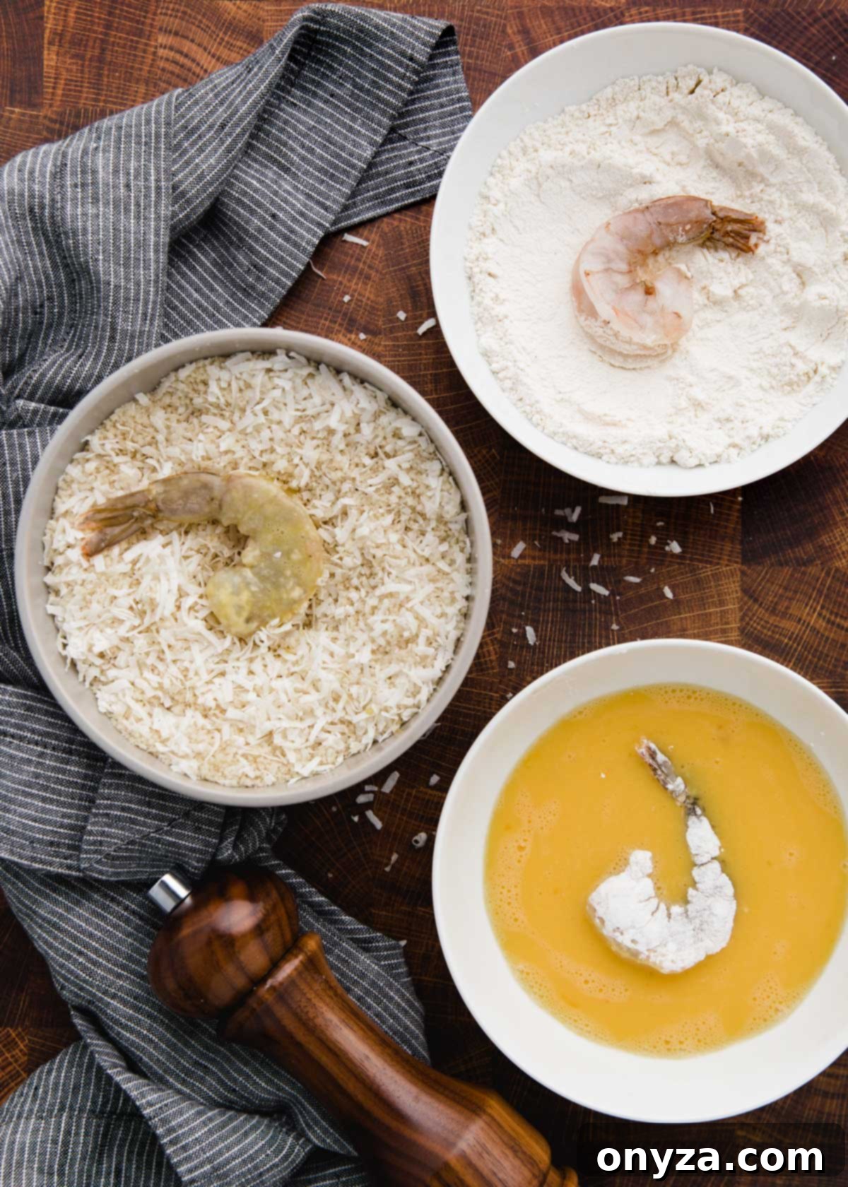 Tropical Crispy Prawns 4 Three shallow bowls set up for breading coconut shrimp, showing shrimp in each stage: flour, egg wash, and the coconut-panko mixture.