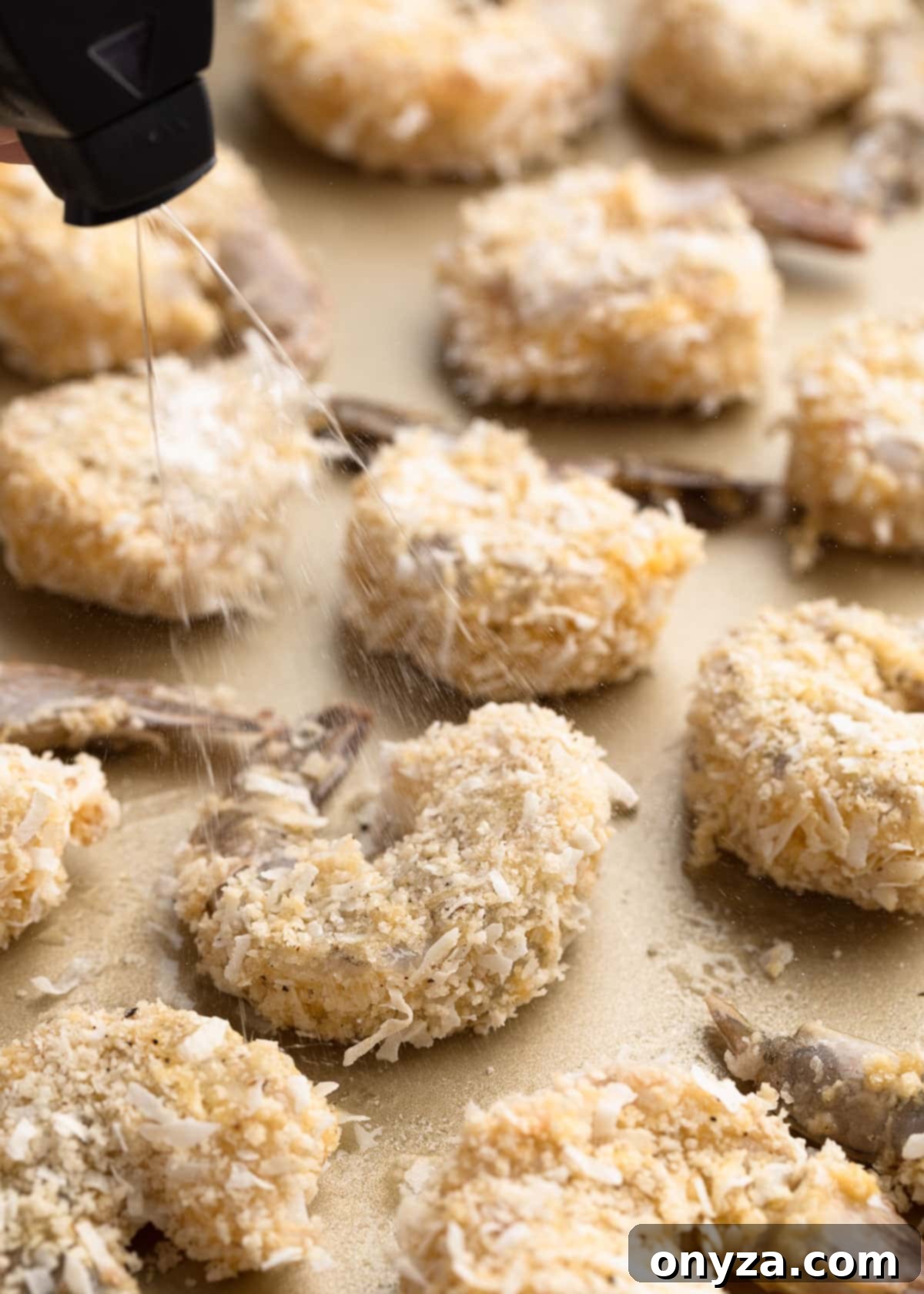 Tropical Crispy Prawns 9 Raw coconut shrimp on a baking sheet, being sprayed with cooking oil from a mister bottle before baking.