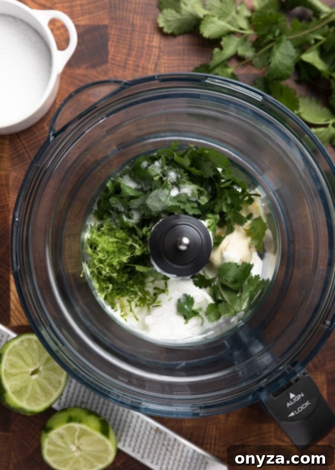A food processor bowl filled with fresh ingredients including vibrant green cilantro leaves, bright yellow lime zest, creamy mayonnaise, and rich sour cream, all ready to be blended into a delicious sauce.