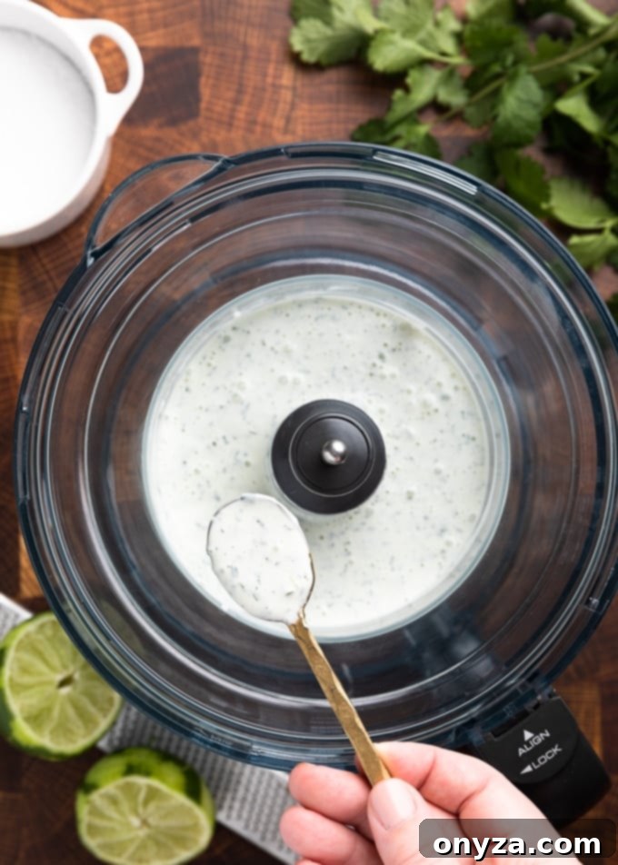 A close-up shot of a spoon filled with creamy, vibrant green cilantro lime sauce, held over a food processor bowl, showcasing its smooth texture and rich color.