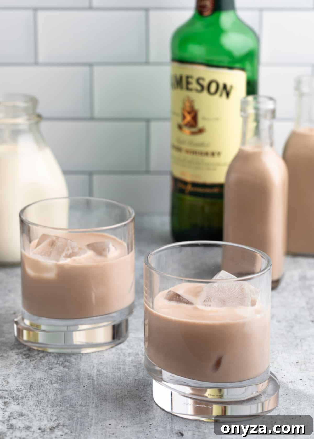 Two glasses of homemade Irish Cream with ice, set against a backdrop of small liqueur bottles, a bottle of Irish whiskey, and a carton of heavy cream, symbolizing freshness and homemade quality.