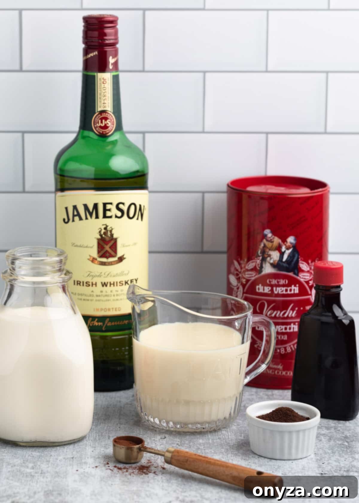 A collection of essential ingredients for making homemade Irish Cream, including bottles of Irish whiskey and heavy cream, jars of sweetened condensed milk, cocoa, instant coffee, and vanilla extract, arranged on a counter against a white tile backsplash.