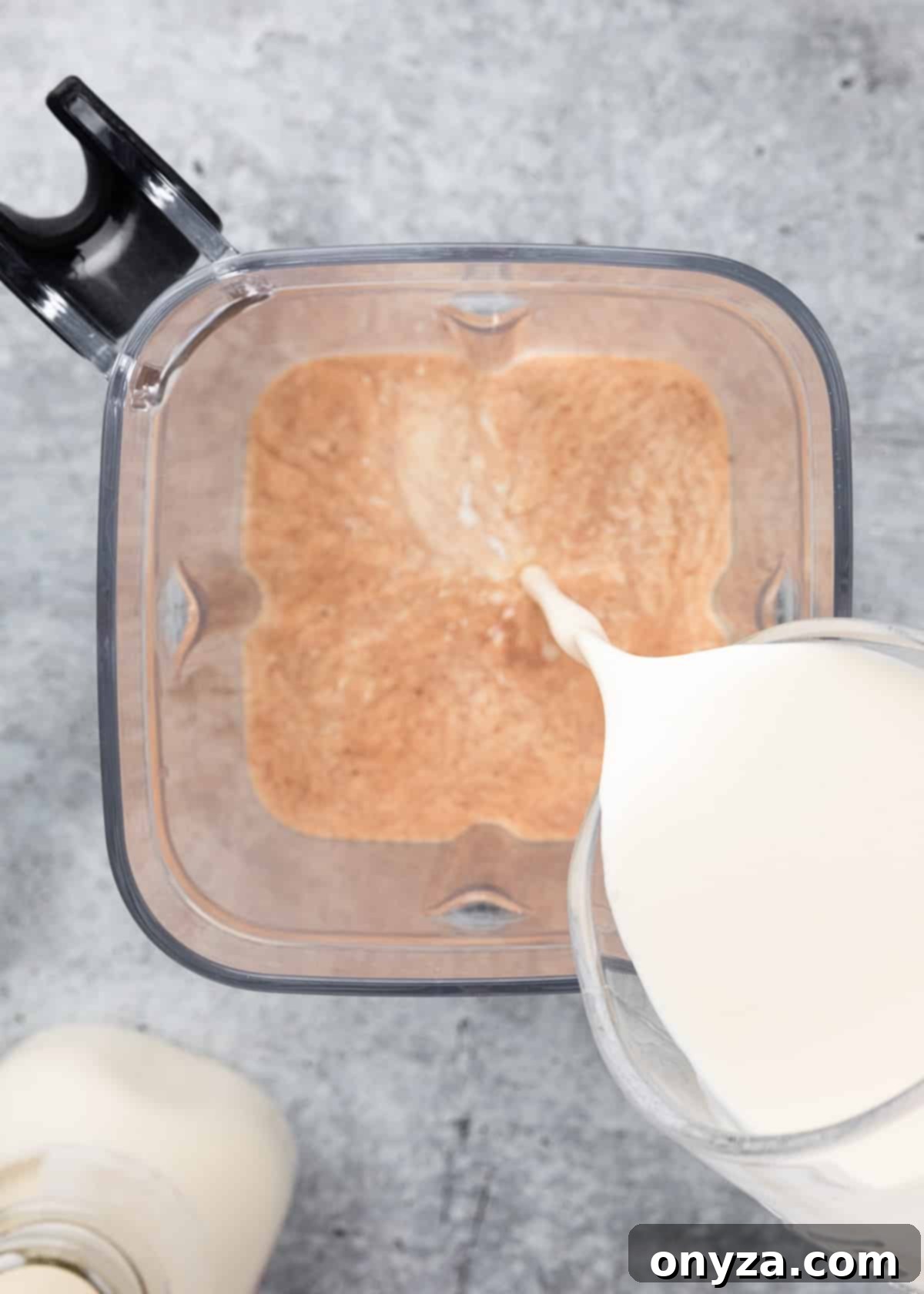 Fresh cream being poured into the partially blended homemade Irish Cream mixture in a blender canister, marking the final stage of preparation for a smooth liqueur.
