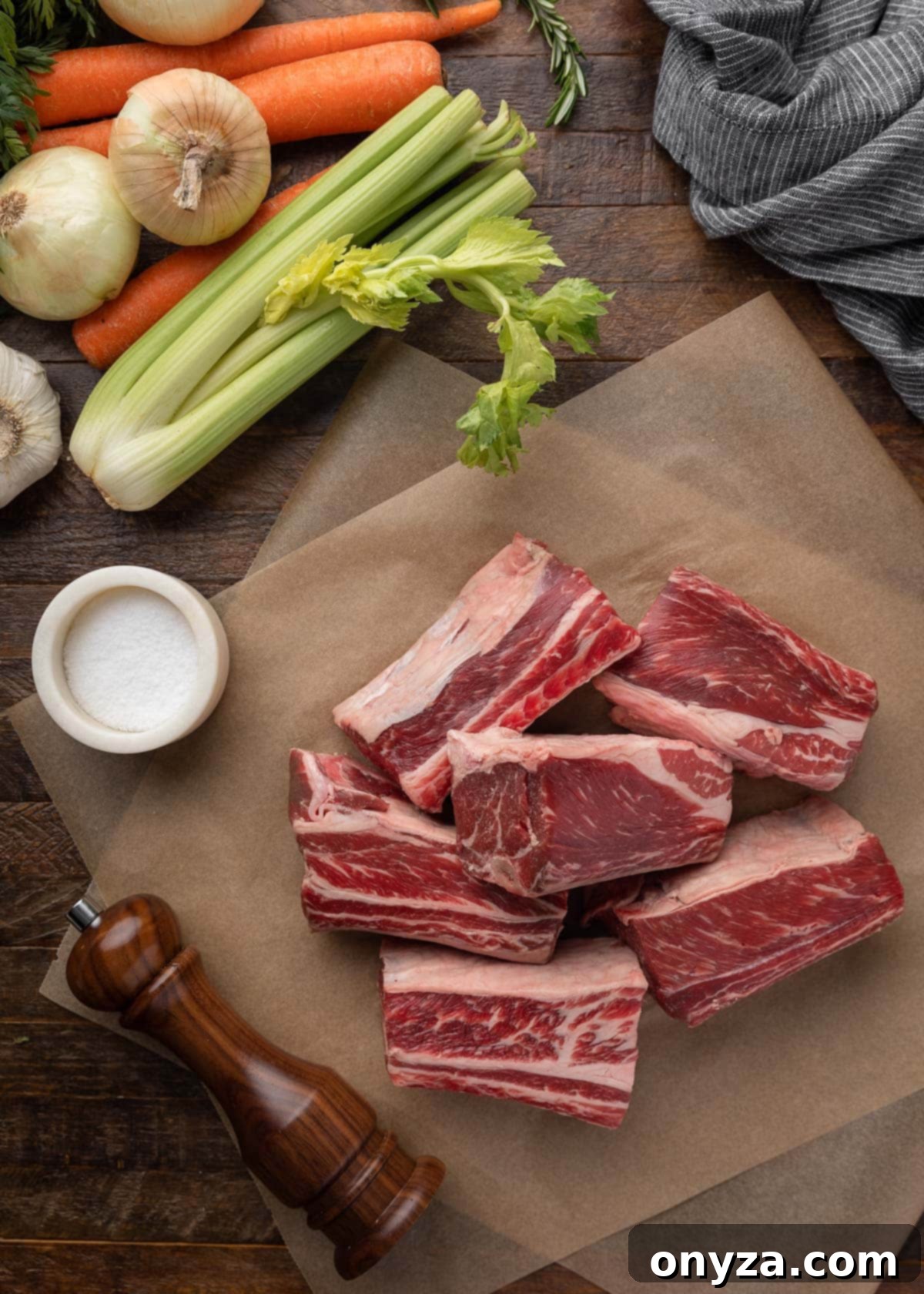 Raw short ribs on parchment paper, surrounded by celery, onions, carrots, a salt crock, and wood pepper grinder – key ingredients for braising