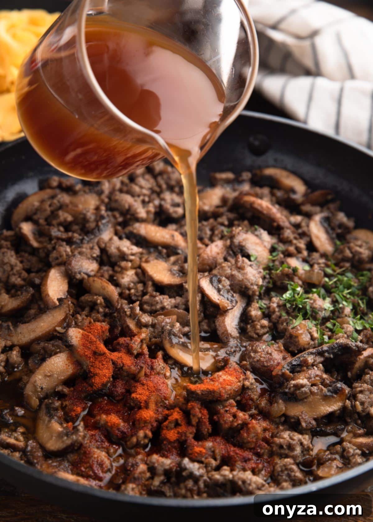 pouring beef broth into a saute pan with ground beef, mushrooms, paprika, and herbs