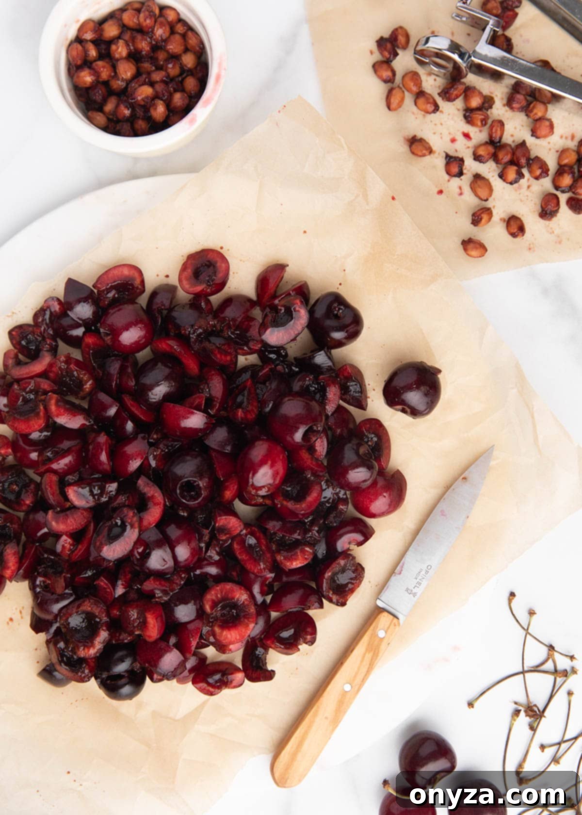 Chopped cherries on a parchment-lined marble board, with a paring knife, cherry pitting tool, and bowl of cherry pits.
