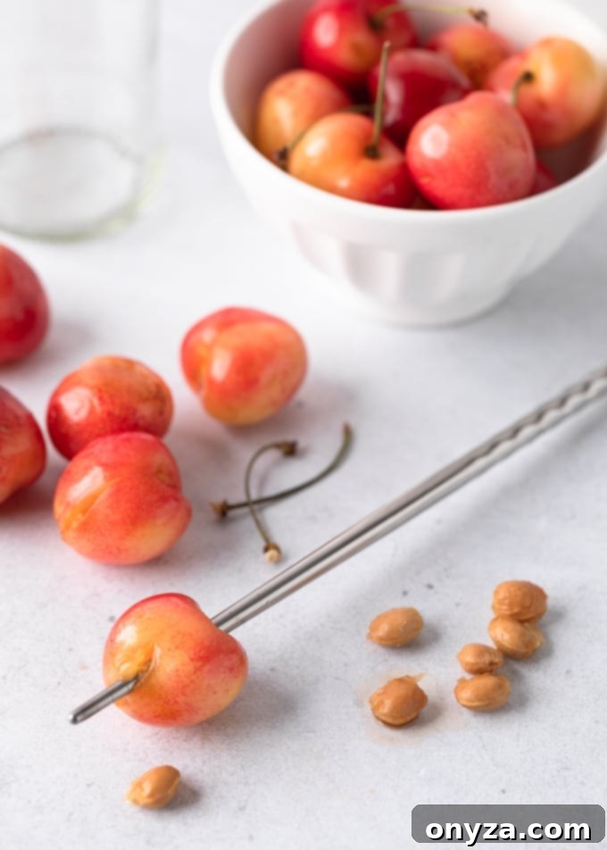 A Rainier cherry skewered with a metal chopstick next to a pile of cherry pits, pitted cherries, and a bowl of whole cherries