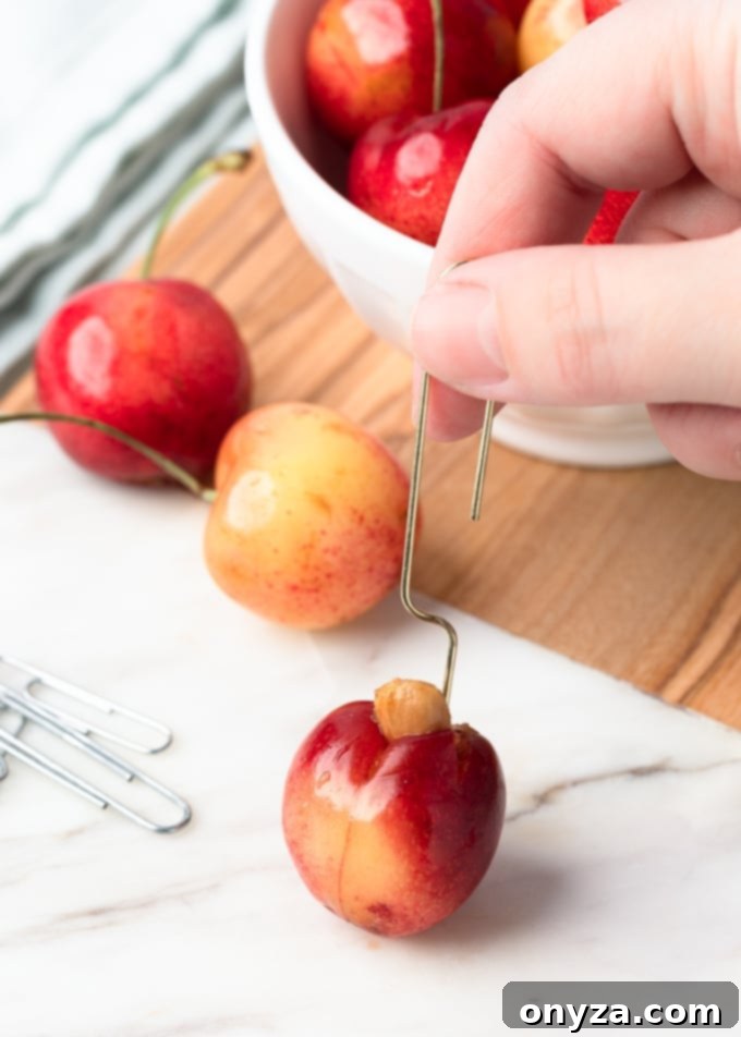 Pit being removed from a Rainier cherry with a gold-tone paper clip, on a marble cutting board