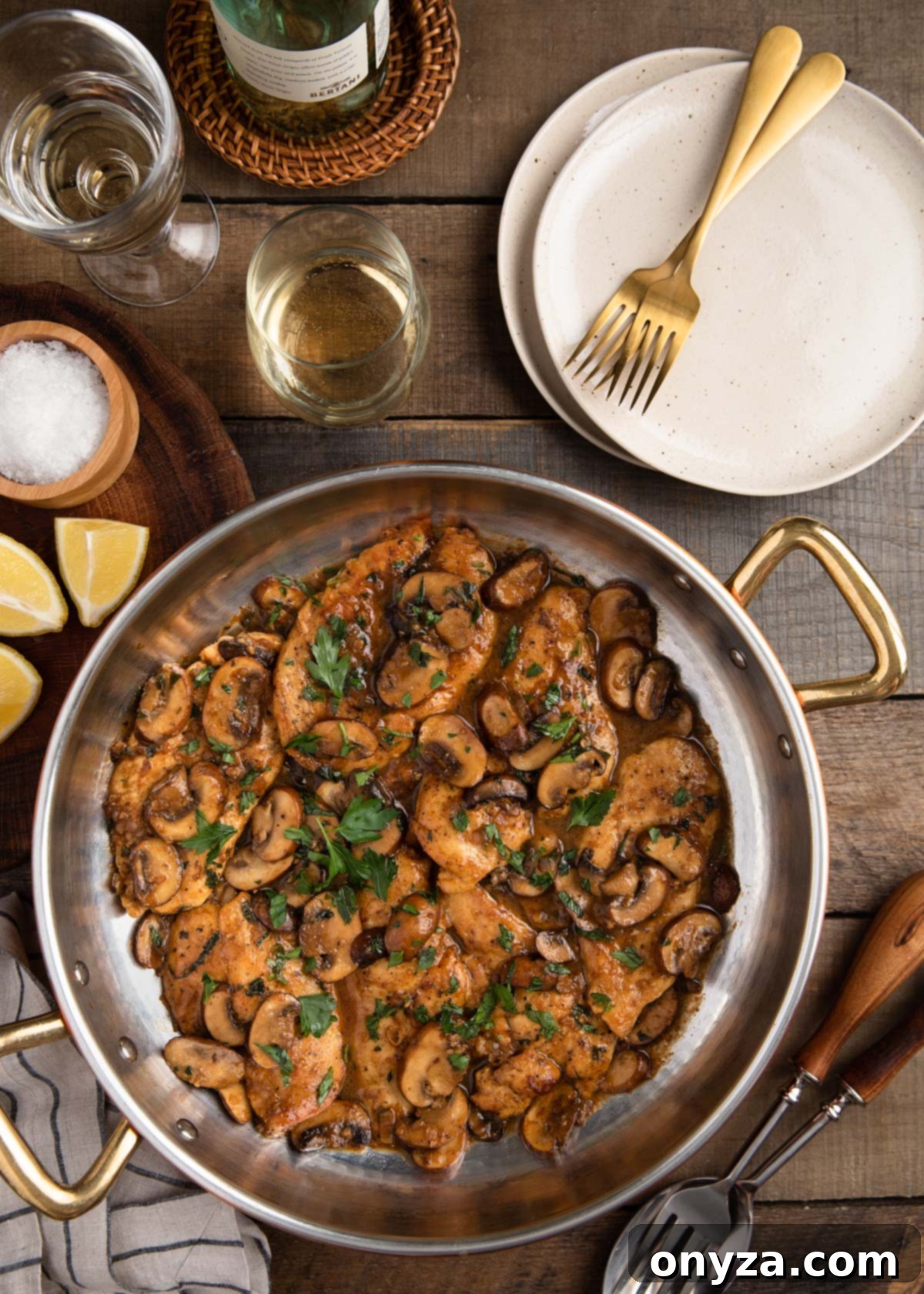 Overhead shot of perfectly cooked Chicken Marsala in a gleaming copper pan, accompanied by serving plates and wine glasses, all set on a rustic wooden board.