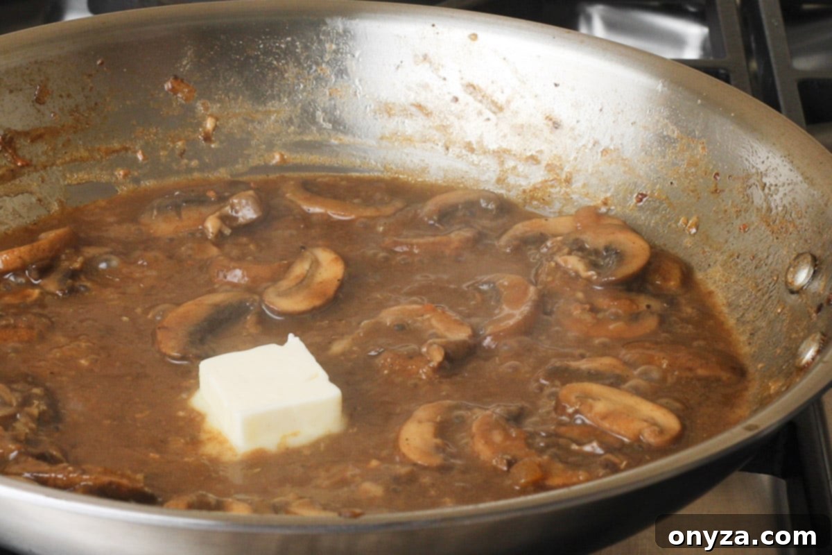 A knob of unsalted butter being swirled into the finished Marsala sauce in a stainless steel frying pan, adding richness and gloss.